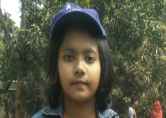 South Asian girl with a denim vest and blue baseball cap standing in front of a leafy tree
