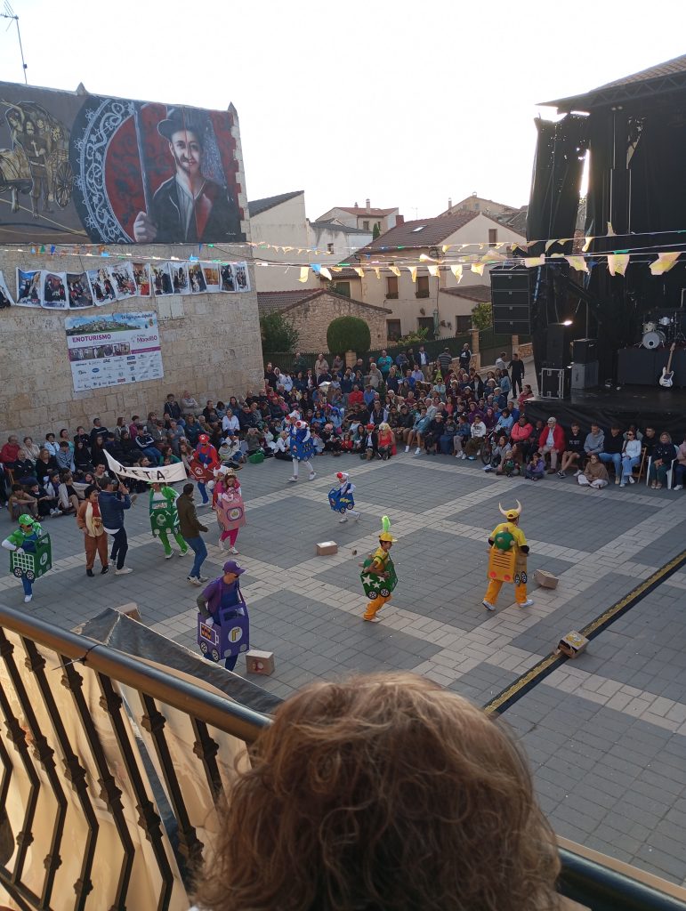 People in yellow, green, blue and red outfits down in a concrete town square dancing and showing off. Spectators watching from a balcony. 