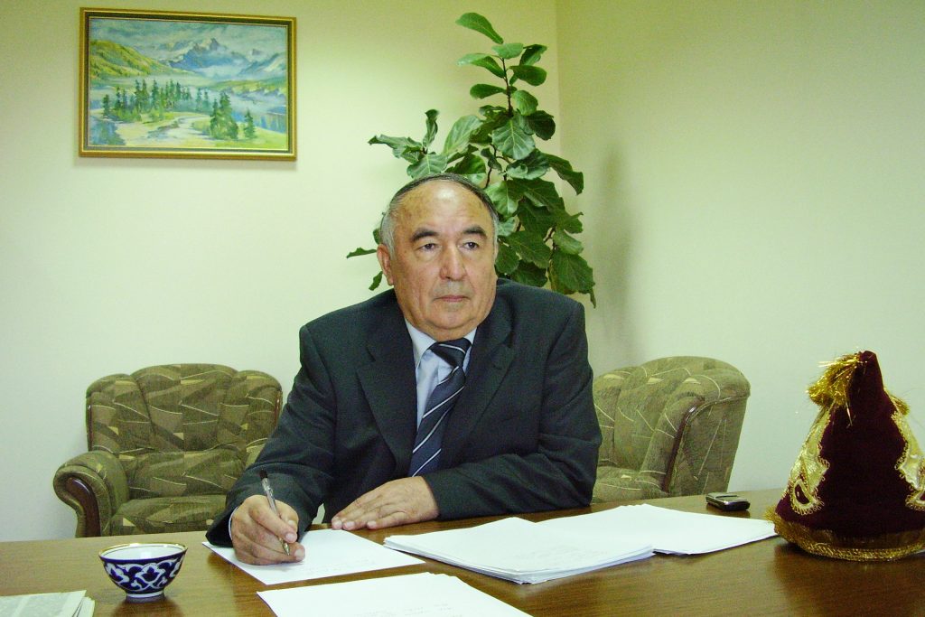 Older light skinned Central Asian man with a balding head, a black coat, white shirt and tie. He's seated at a desk with a plant behind him and a painting of a mountain scene on the wall. He's got paper on his wooden desk where he sits and is writing with a pen. 