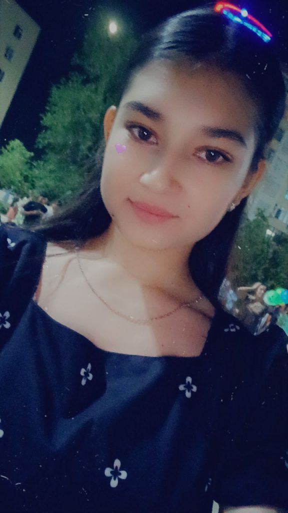 Central Asian teen girl with long dark hair and brown eyes and a small necklace. She's wearing a dark floral blouse and is in front of trees and people at night. 