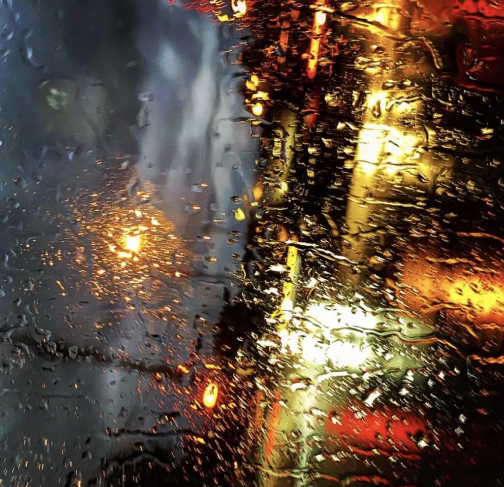 Sideways image of raindrops on a window highlighting gray pavement and white and orange and red lights of buildings and cars ahead. 