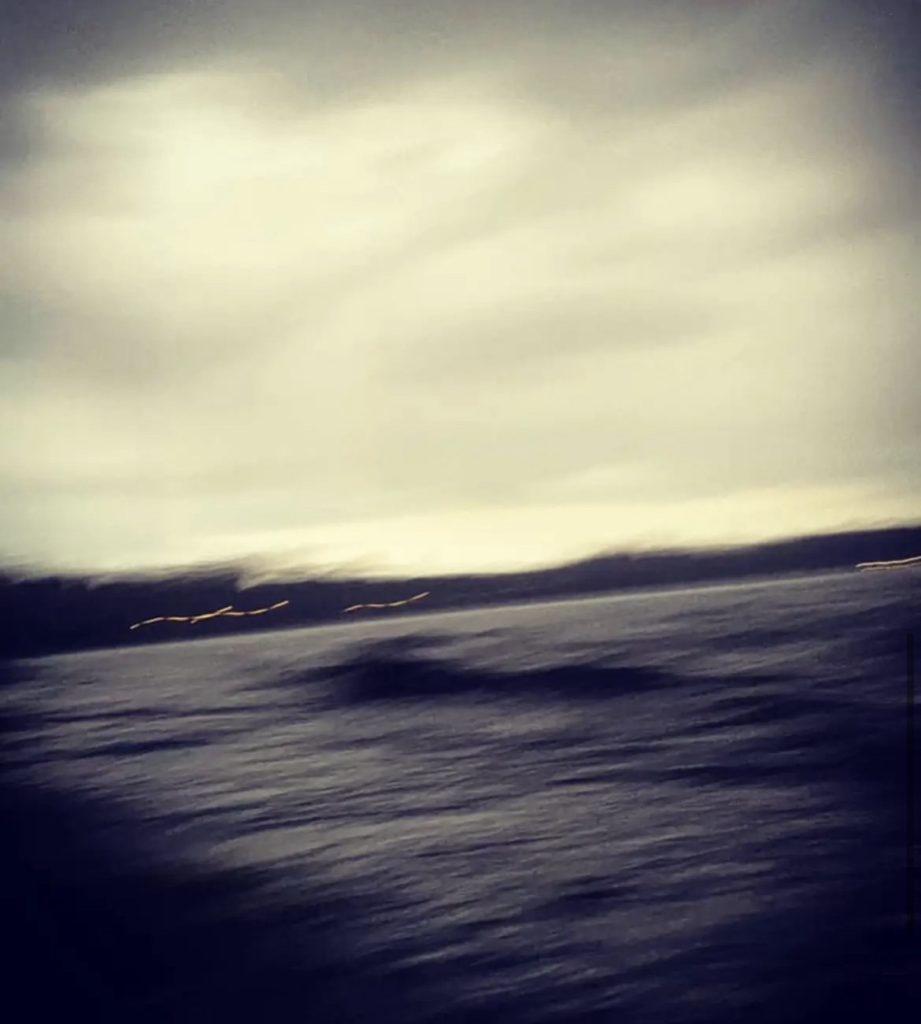 Blurry image of moving ocean water with clouds in the distance and a grey sky. 