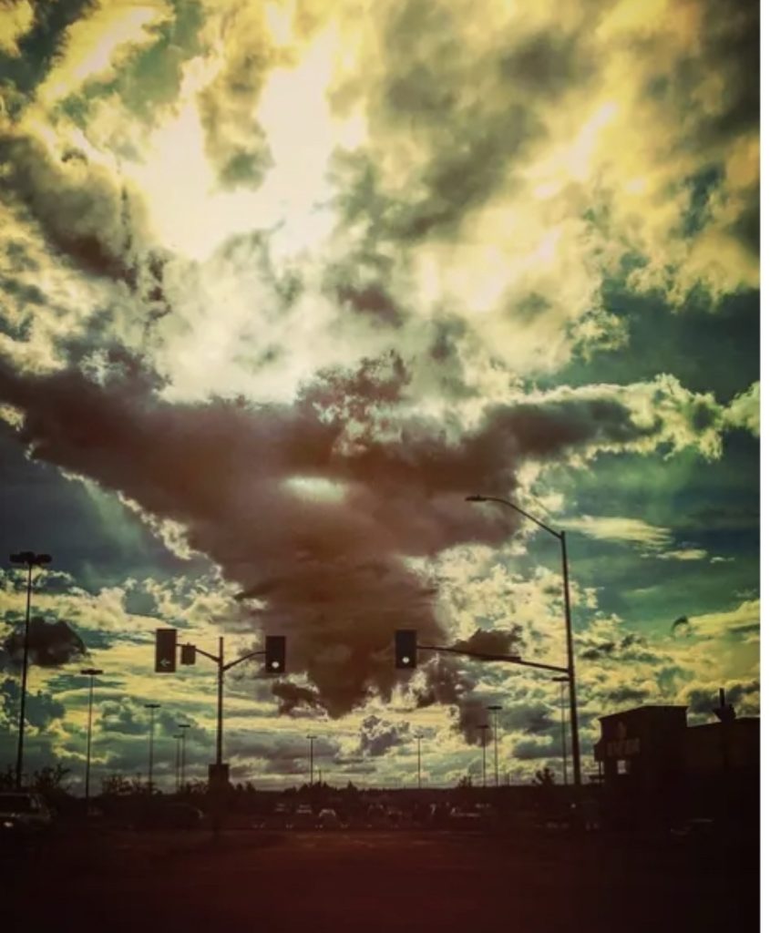 Huge mass of clouds covering the sun and blue sky peeking through above two green streetlights. Everything below is hazy. 