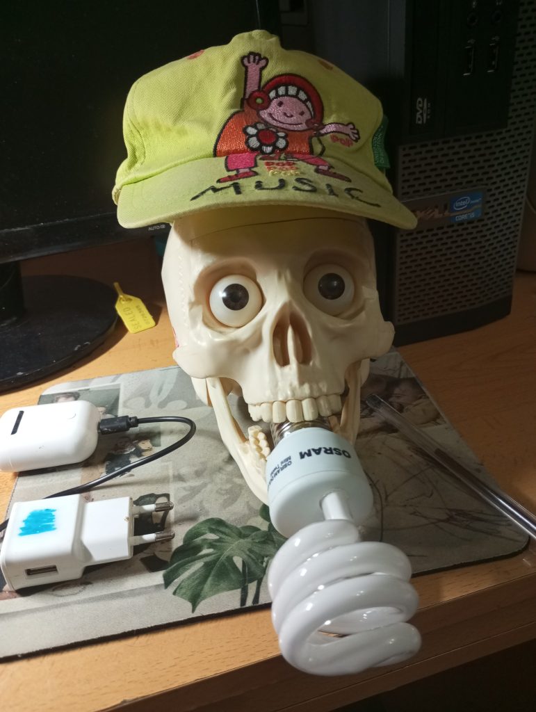 Fake skull with fake eyeballs with a lightbulb in its mouth with a yellow hat that says "Music" on it and has a dancing girl with flowers on her outfit embroidered onto the hat.
