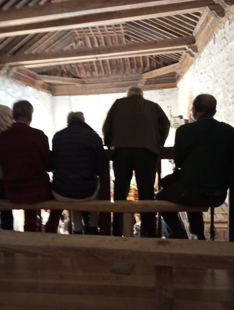 The backs of four seated old men in the stone church's side chapel. Wooden ceiling. 