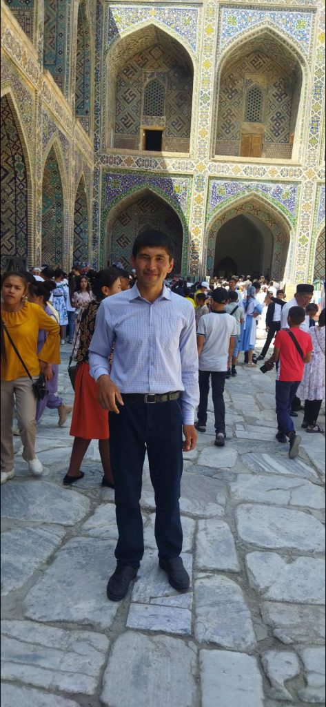 Central Asian man standing in front of an ancient building with Islamic architecture, mosaic designs and sloping arches. He's in a collared shirt and jeans with a belt and in a crowd of people. 