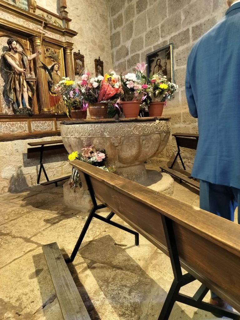 Pots of flowers inside the stone baptismal font of an old stone church with wooden pews and statues of Mary and Jesus. 