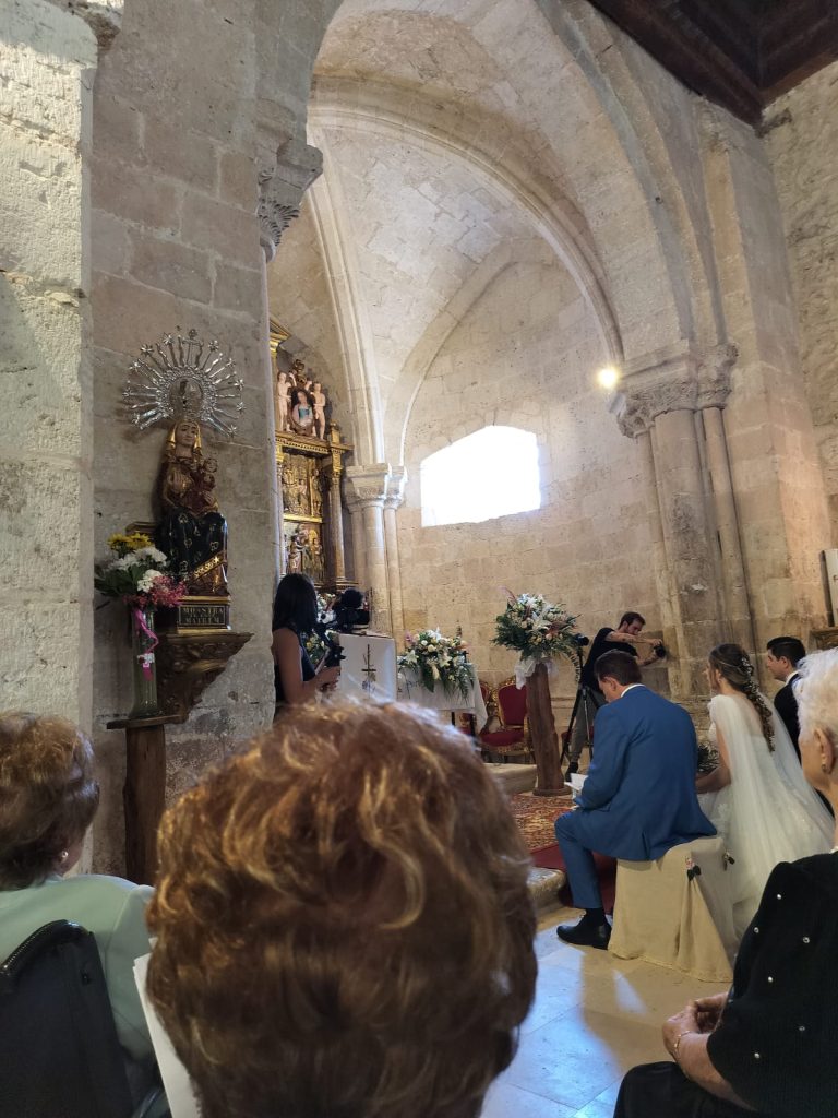 Old stone church with archways and columns and people gathered in a side chapel praying in front of an altar. We see the bride and several well dressed guests. 