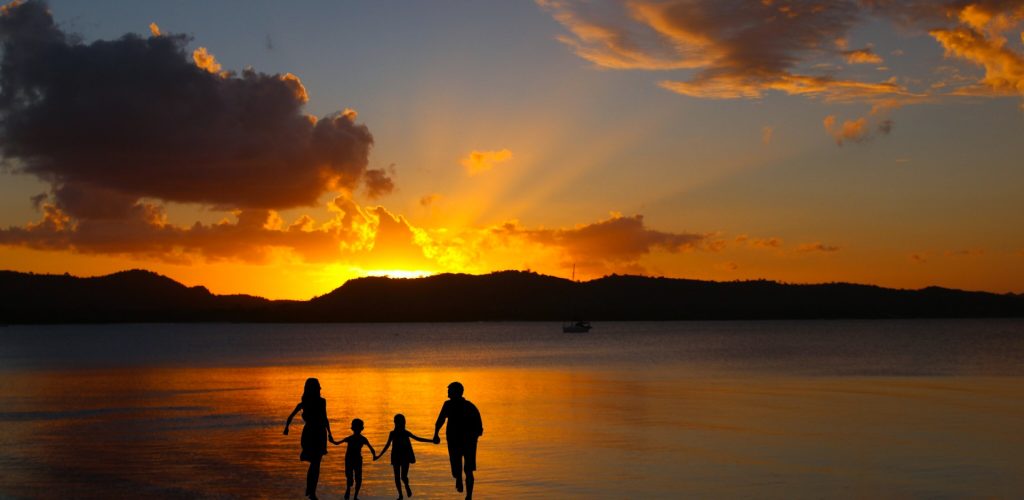 Silhouetted family, two adults and two kids, on a beach at sunset. Mountains across the lake. 
