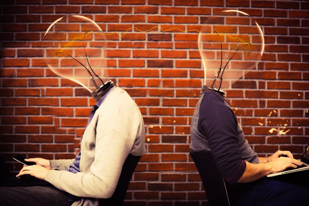 Two light skinned people using laptops at desks not facing each other. Both have lightbulbs for heads and electricity is passing between them. Brick wall in the background. 