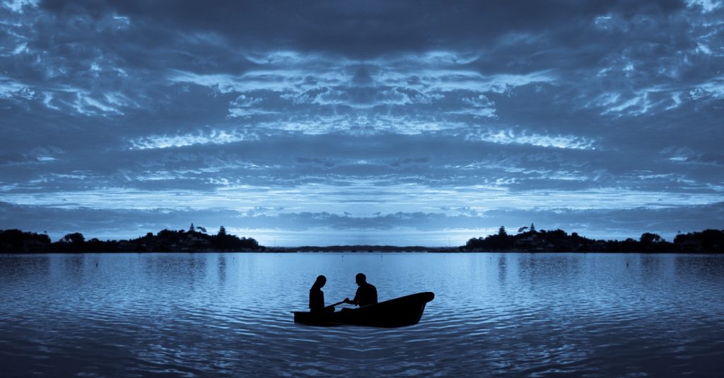 Two figures in a boat with oars off in a lake at sunrise or twilight. Land off in the distance, everything is blue or shadowy black. 