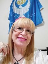 Light-skinned woman with light blond hair and pink lipstick and a pink hair bow and reading glasses. Blue and white peace flag with a dove behind her. She's got a fluffy white blouse and a beaded necklace.