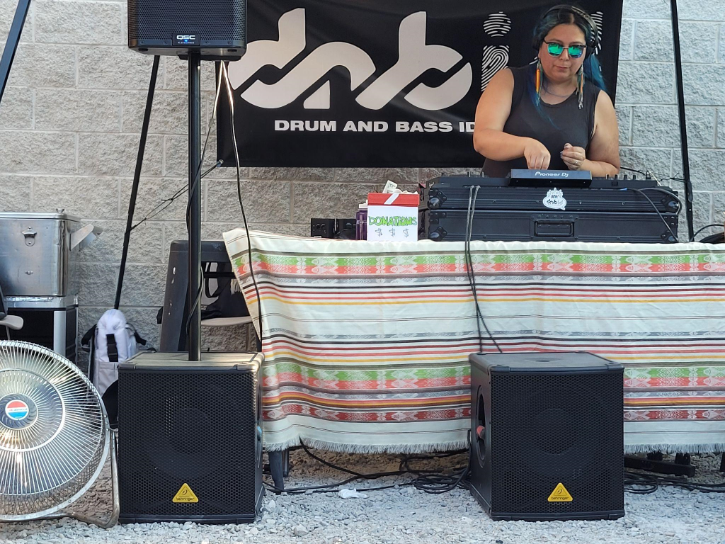 DJ Toyacoyah, a brown skinned person with sunglasses and a black tank top, plays music from a DJ booth with a multicolored cloth and speakers nearby. 