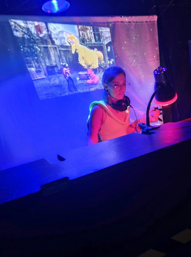 Blonde white woman with her hair in a braid and reading glasses sits at a desk with a video screen with a scene from Sesame Street with Big Bird behind her. She's got earphones on her neck and a lamp on and is producing electronic music! 