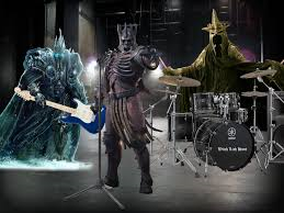 Scary skeleton guy with a loincloth and bare ribs and a crown with two other large caped and crowned monsters in the background. They have an electric guitar, drum set, and cymbals and are in a band. 