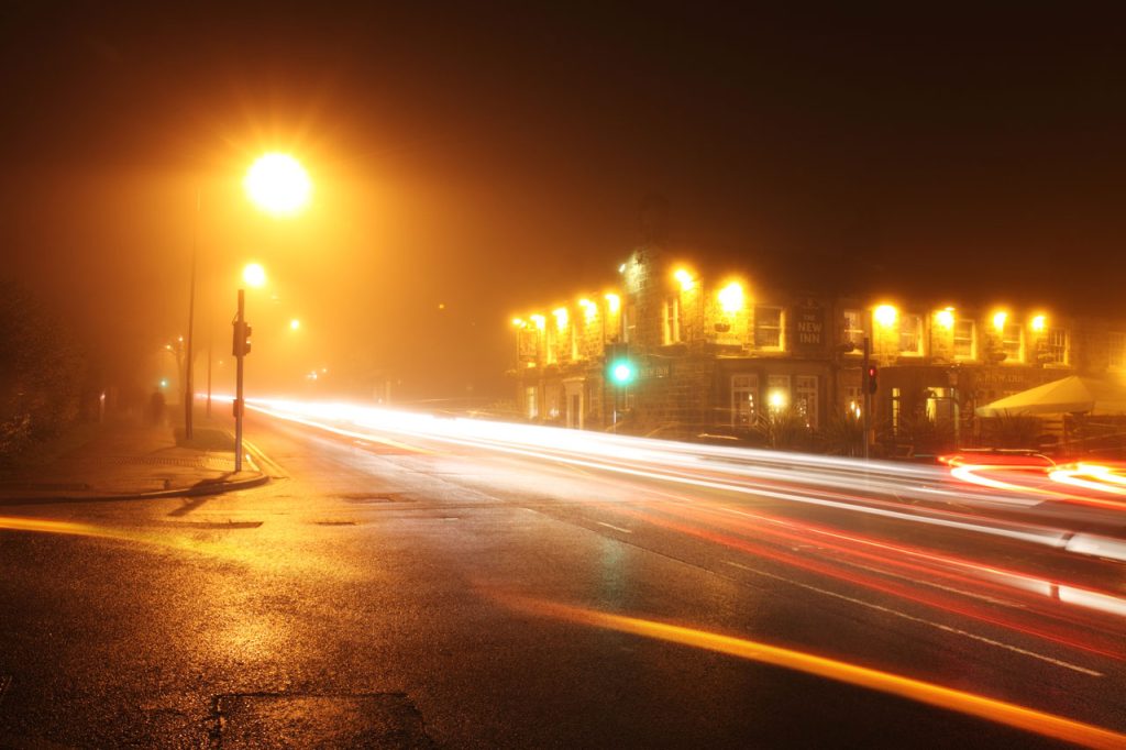 Foggy city street at night with streetlamps and a building with lights on off in the distance. 
