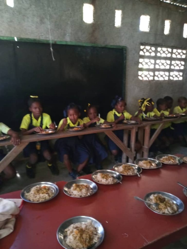 A row of young Black girls in yellow school uniforms sit at a long wooden table and eat a rice dish. 