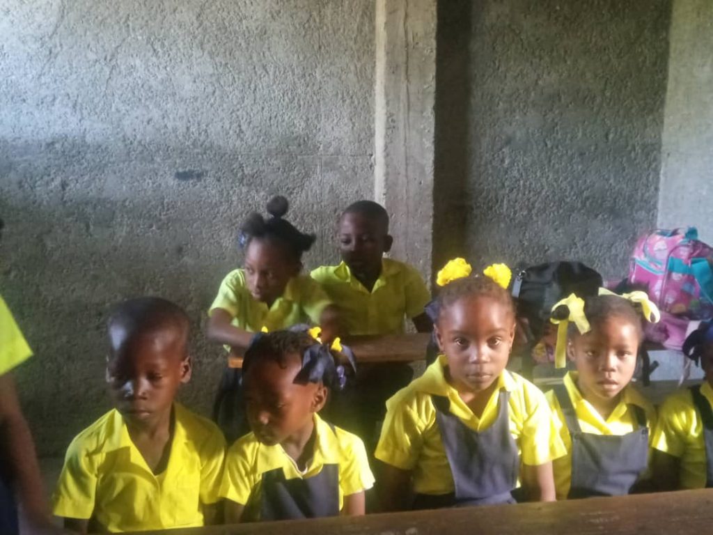 Young Black boys and girls with pigtails and braids and yellow and black school uniforms sit at desks. 
