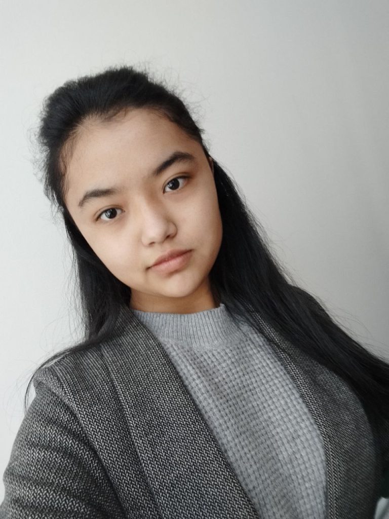 Young Central Asian teen girl with long black hair and brown eyes. She's wearing a gray coat over a gray sweater. 