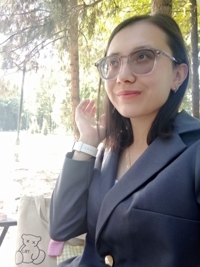 Young Central Asian woman with short black hair, reading glasses, a gray jacket and necklace and white wristwatch. She's outside near some green conifer trees. 