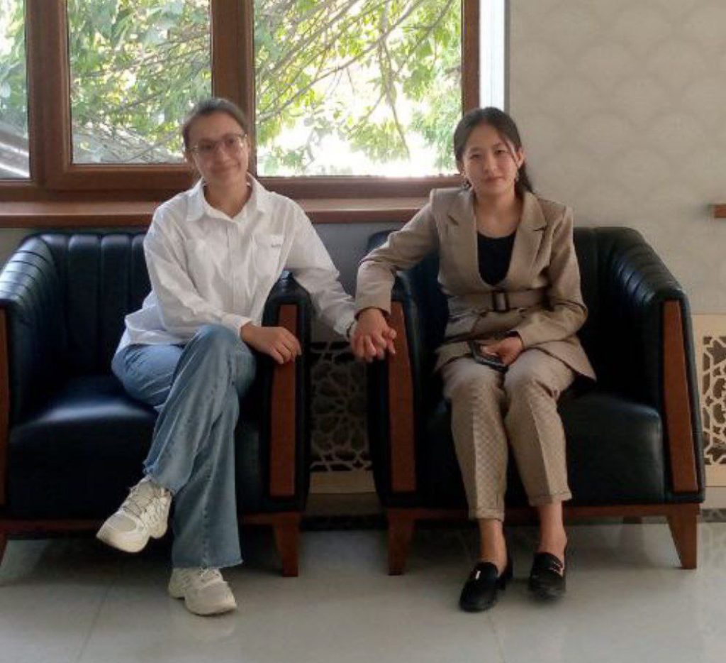 The author, Bakhora Bakhtiyorova, sits in a poofy black chair on the left in a white blouse and blue jeans. Her sister holds her hand and sits next to her and is dressed up in a brown business suit. There are pine trees out the window.