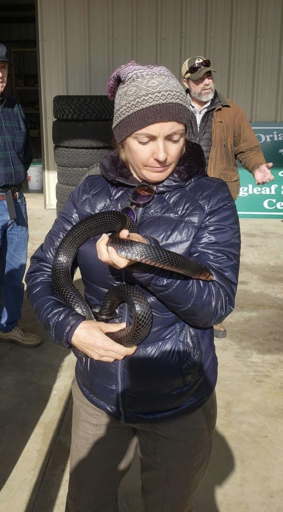 Young white woman holding a dark scaly snake. She's got a knitted hat and a blue puffy jacket. 