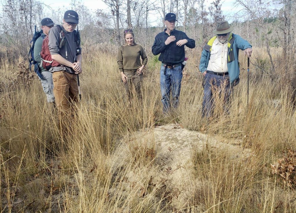 Group of people with jackets and hats on looking at tall grass and dirt and trees outdoors. 