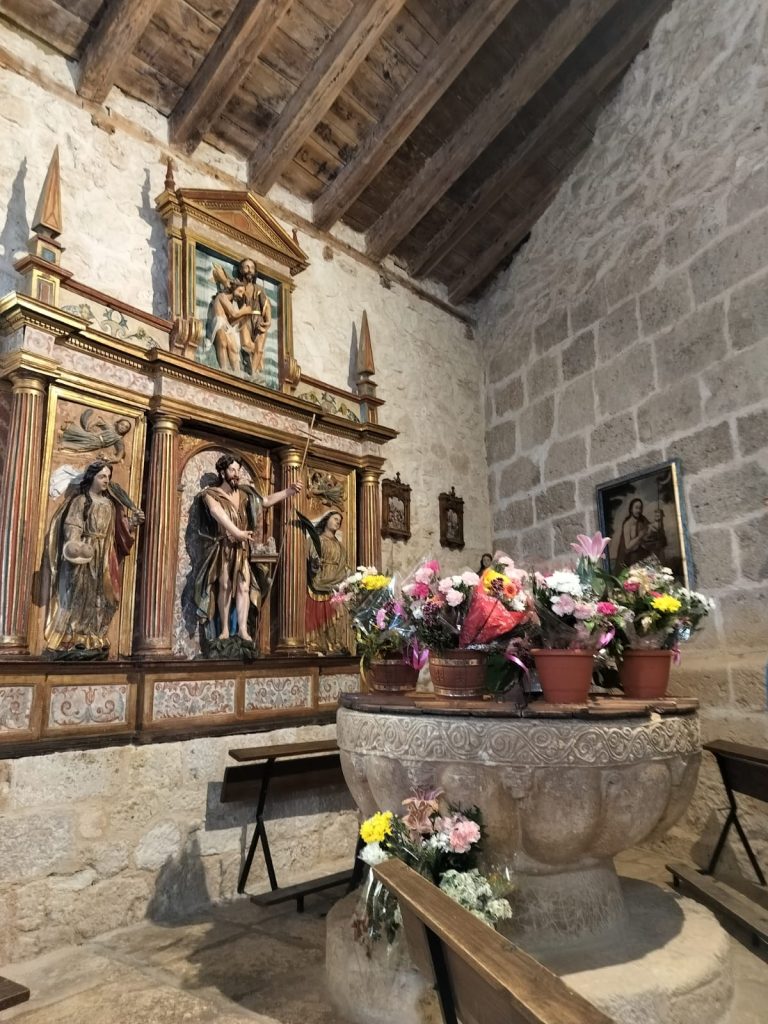 Inside of an old gray brick church with wooden ceilings and an altar with saints and angels in painted wood and potted flowers on the altar. 