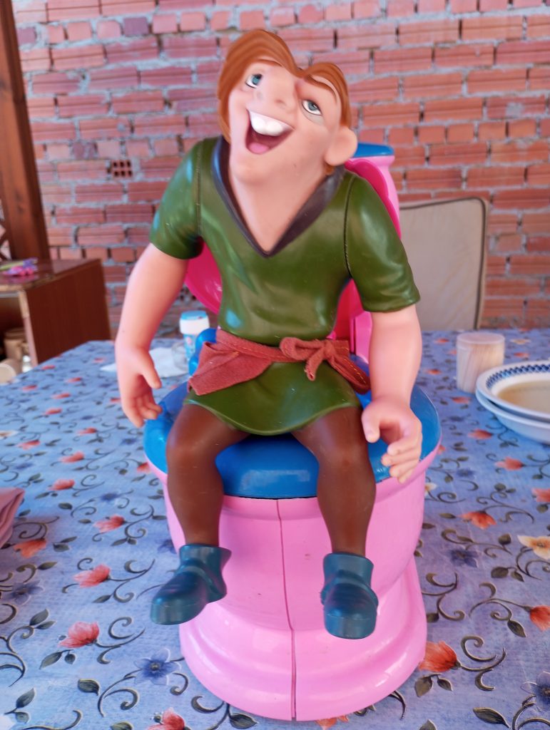 Figurine of a laughing man dressed like Robin Hood with a short dresslike top and a belt. He's on a plastic seat with a table and tablecloth and brick wall behind him. 