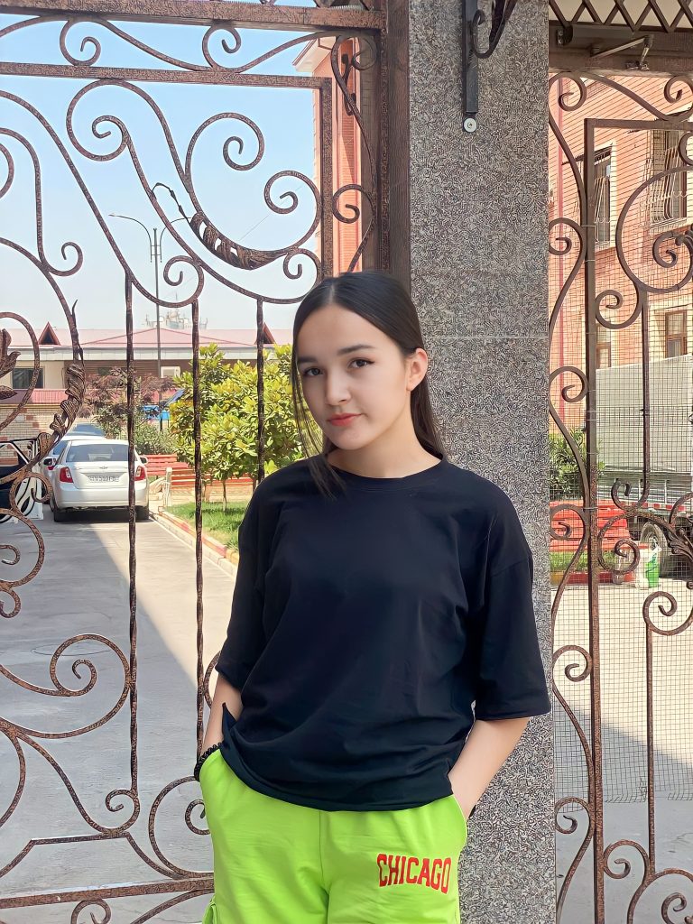 Young Central Asian teen girl with dark straight black hair and brown eyes. She's got a black tee shirt and bright green pants and is in front of a metal gate with a street and a car and an apartment building in the background. 