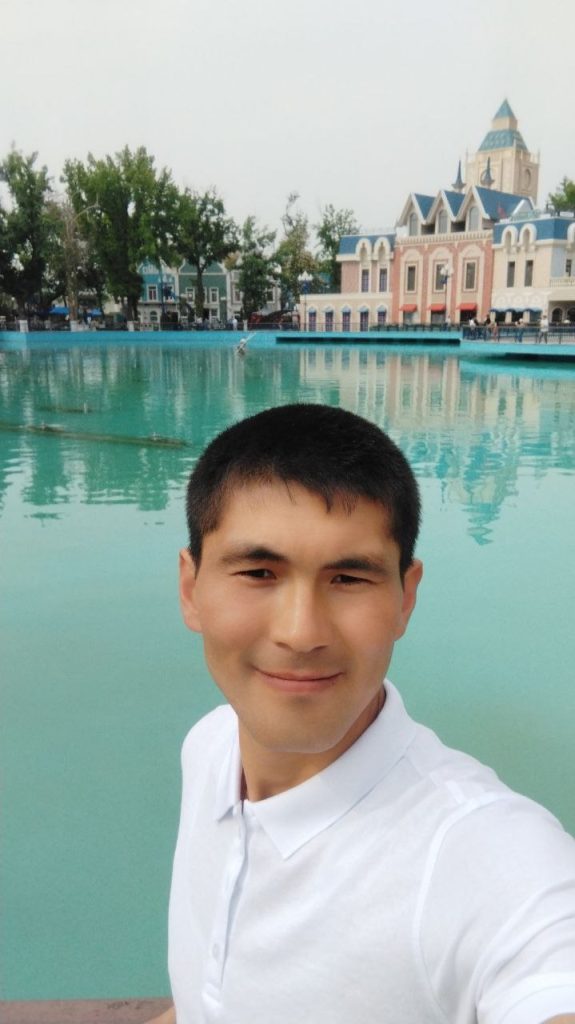 Young Central Asian man with a white collared shirt and short brown hair standing in front of a pool in a park. Castle-like building and trees on the other side of the pool. 