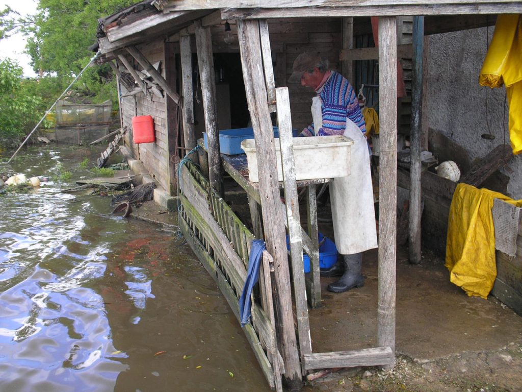 Older fisherman in a striped sweater and hat in a small wooden shelter by the side of a lake with some trees. Poles are in the water. 