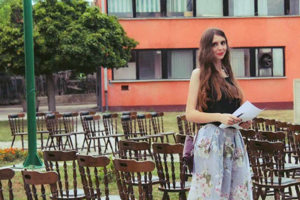 Young light skinned woman with long brown hair, a black top, a blue and white floral patterned skirt standing amidst a bunch of wooden chairs outside on a lawn. There's a lamppost and a building nearby.