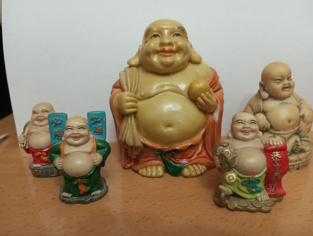 Large fat ceramic statue of Buddha flanked by two smaller Buddhas on each side. Some hold apples or signs with Asian language writing. They have robes and trousers. 