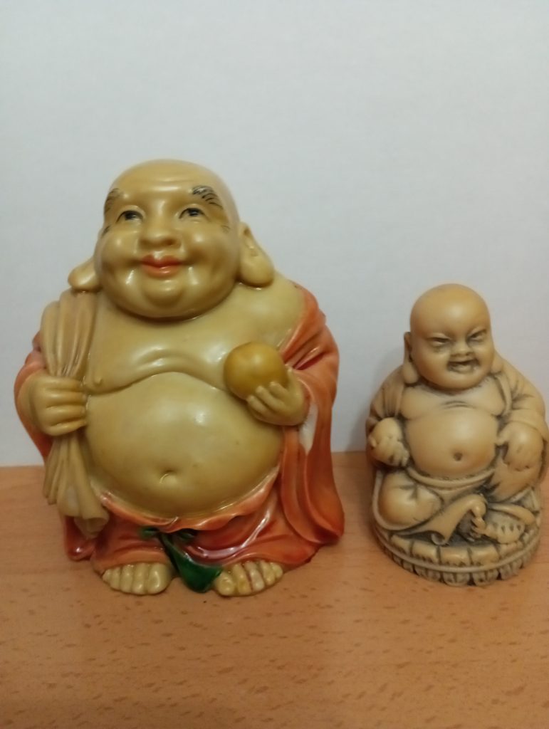 Two Buddha statues, the one on the left is larger and holds an apple and wears a red robe while the one on the right is smaller and a solid brown. 