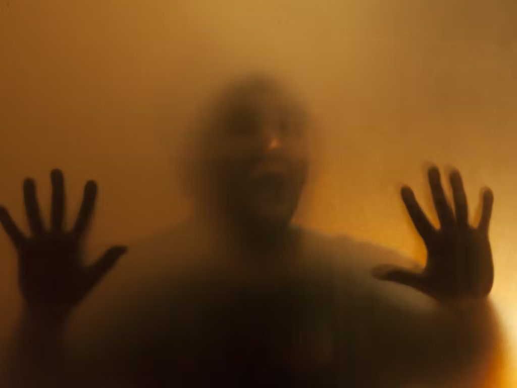 Middle aged man of indeterminate race behind translucent glass holding up his hands to his side and against the glass. 