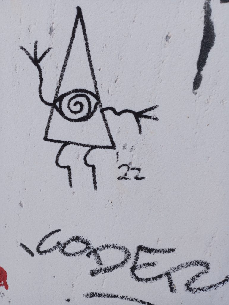Line drawings of black pen or chalk on a white concrete surface. Triangle with an eye and legs and arms. 