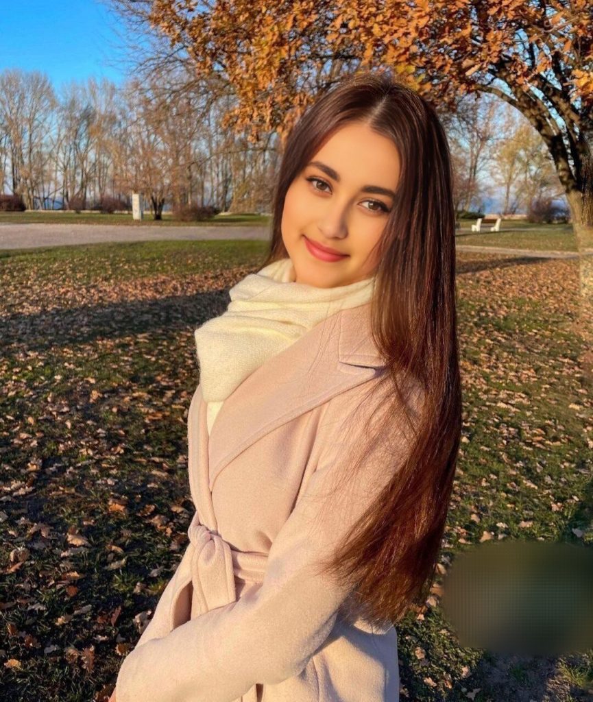 Young Central Asian woman with long dark hair, a white scarf and a pink coat standing outside in a grassy field under a tree with orange leaves. 