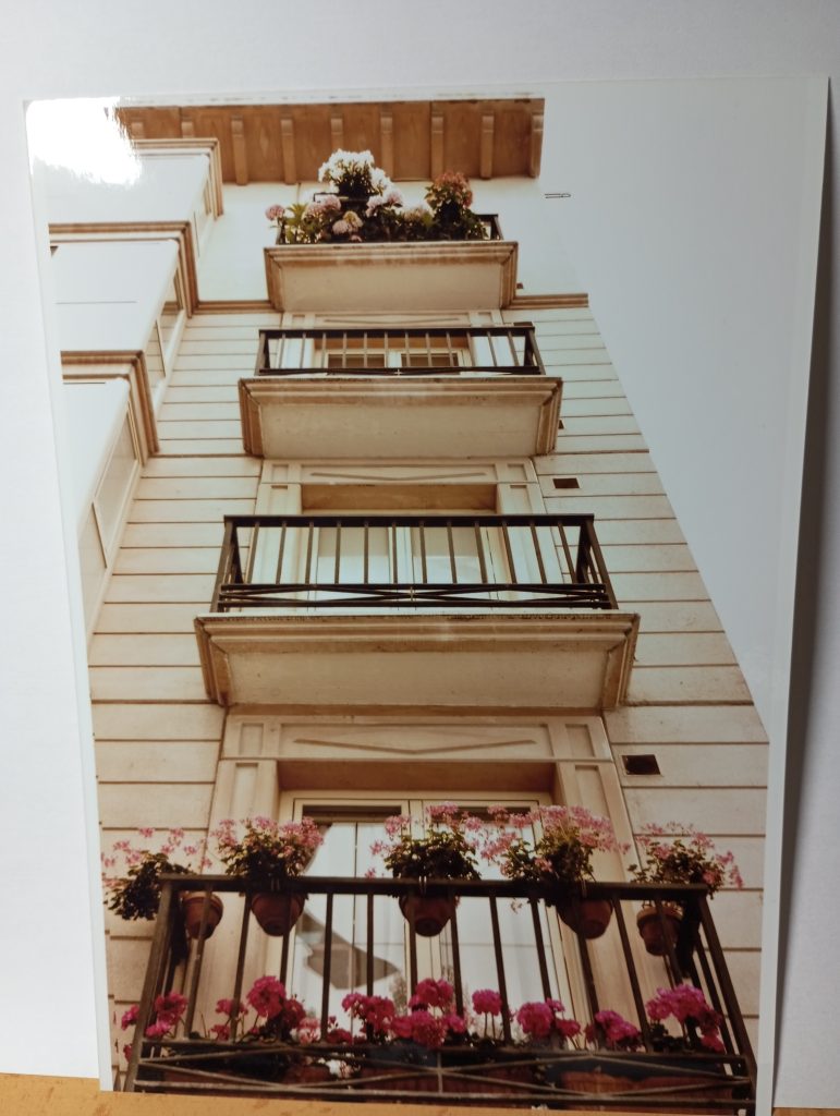 Off white building photographed from the bottom, four balconies, the top and bottom have pink flowers. 