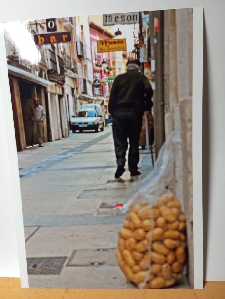 Old man with a coat and dark jeans walking down a street in Europe with small restaurants, a car, and a sack of potatoes. 