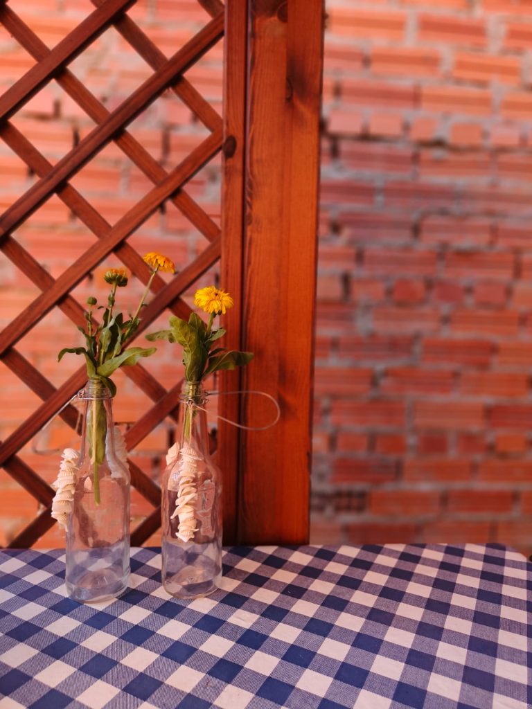 Dandelions in bottles on a blue and white checkered tablecloth with a wood fence and a brick wall in the background.