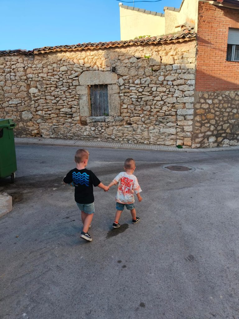 Two young boys in tee shirts and tennis shoes and shorts walking on a gravel street towards an old stone building with a window. 