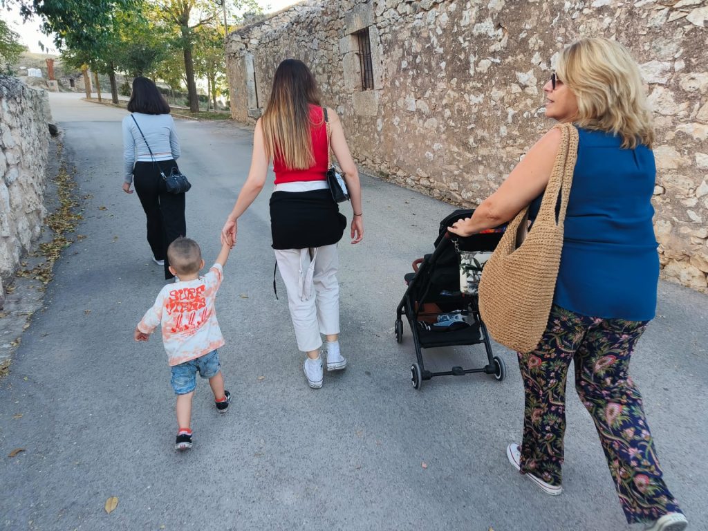 Three middle aged women seen from behind, dressed in short-sleeved street clothes, walking. One has a child by the hand, another pushes a stroller. They are on a narrow street between old stone buildings. 
