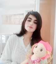 Young light skinned teen girl with long straight dark hair, brown eyes, makeup, and a light white blouse. She's standing in front of a window and holding a doll with a pink hat and outfit.