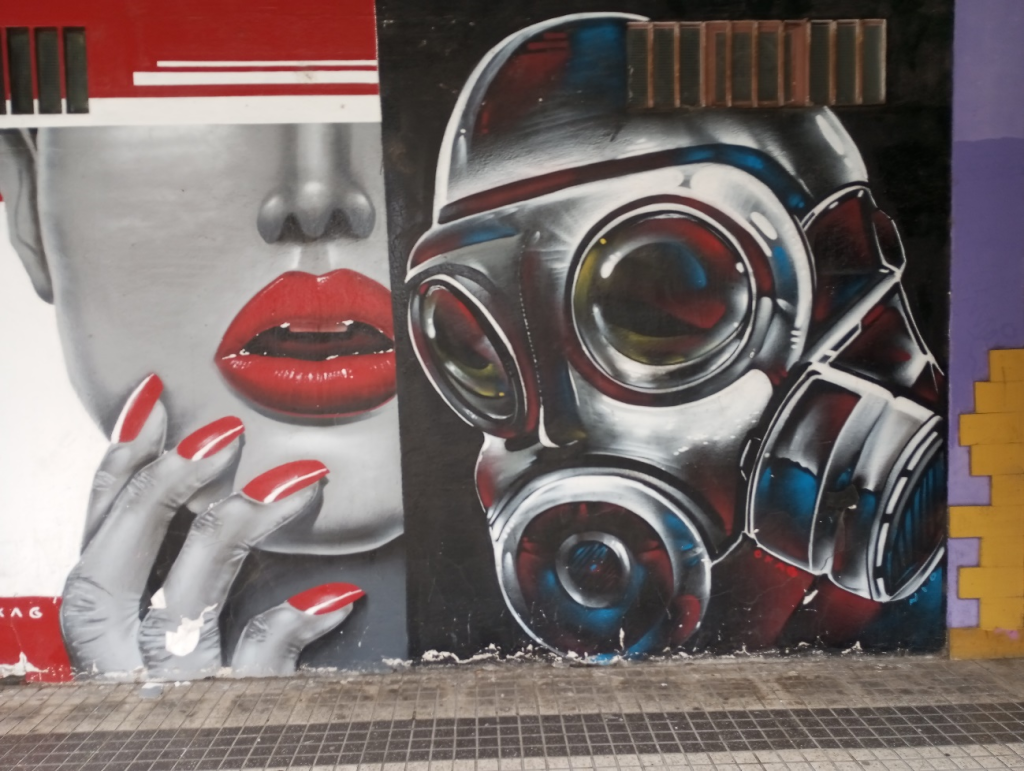 Paintings next to each other on the sidewalk propped against a purple and yellow wall. One of a woman's hands and face with red lipstick and nail polish and another of a gas mask on a black background. 