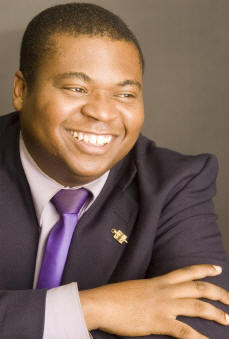 Young Black man, smiling, with short hair on the top of his head. He's wearing a suit and purple tie. 