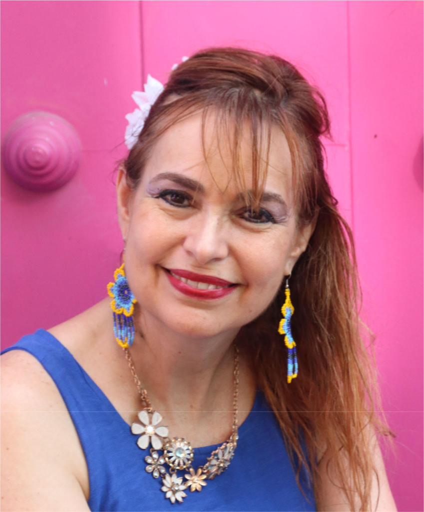 Latina woman with reddish-brown straight hair, lipstick and eyeshadow, blue and yellow earrings, a large floral necklace, and a blue tank top, with a pink wall behind her. 