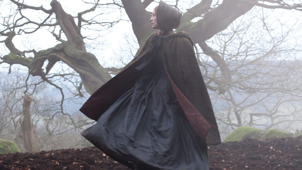 Young white woman with a thick cape and long blue dress waving in the wind outside in the fog near a tree empty of leaves. 