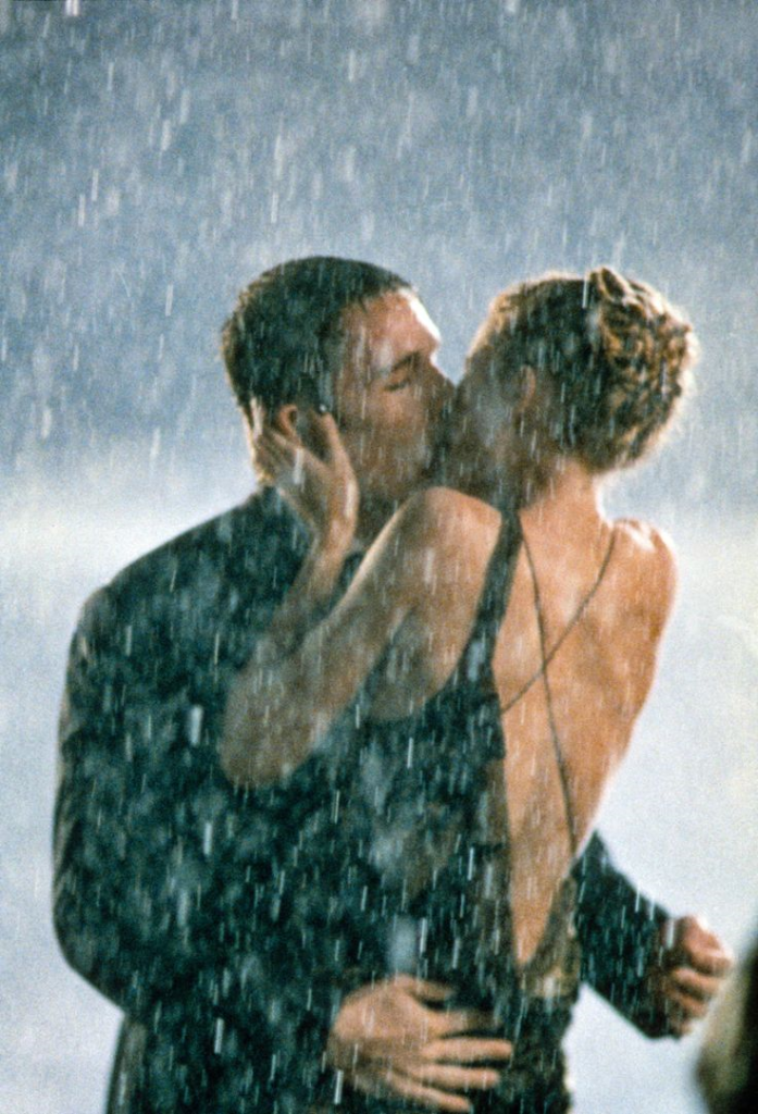 Two light skinned people, a man with a dark jacket and a woman with a black dress falling over her shoulder, kissing in the rain. 