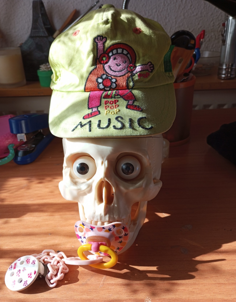 Skull on a wood floor near toys with a pacifier in its mouth and a yellow baseball cap with a clown and the words Pop, Pop, Pop Music in orange and pink and yellow. 